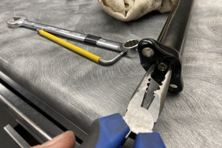 Image showing a set of tools on a metal work surface. Included are a pair of pliers, a wrench, a yellow-handled tool, and a fabric tool bag. The tools are in various orientations, with the focus on the pliers gripping a black component.