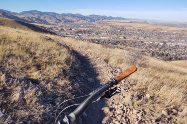 A view of a mountain biking trail on a hillside, with a focus on the bike handlebars in the foreground. The landscape features rolling hills and a valley below, dotted with buildings and vegetation, under a clear blue sky. Green Mountain mountain bike trail.