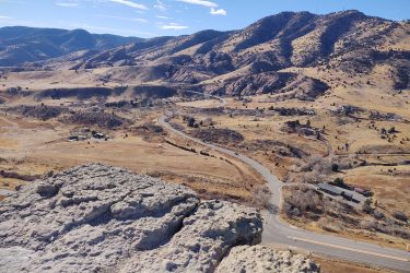 A panoramic view of rolling hills and rugged mountains under a clear blue sky, with winding roads cutting through the golden-brown landscape. Sparse vegetation and a few small structures dot the valley below. Red Rocks / Dakota Ridge mountain bike trail.