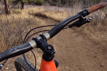 A close-up view of a mountain bike's handlebars overlooking a dirt trail, with a scenic landscape featuring rolling hills and distant mountains under a partly cloudy sky. The surrounding area includes pine trees and dry grass, suggesting a natural outdoor setting ideal for biking. White Ranch mountain bike trail.