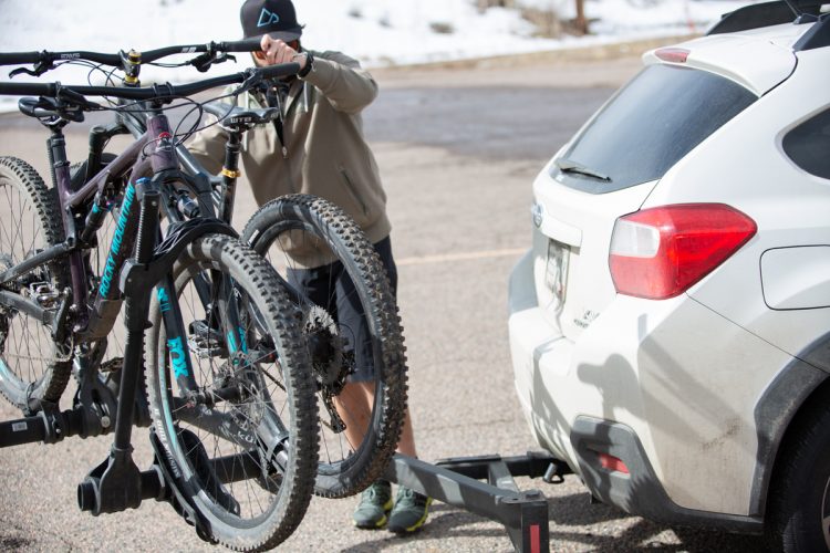 A person is seen using a bike rack to load two mountain bikes onto the back of a white SUV in a parking area. Snowy terrain is visible in the background, suggesting a cold environment. The person is wearing a gray jacket and shorts, with a cap, and is actively securing the bikes before setting off.