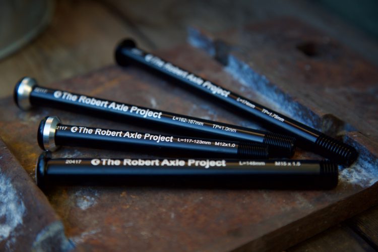 Four black bicycle axles from The Robert Axle Project arranged on a wooden surface, each marked with specifications including length and thread types.
