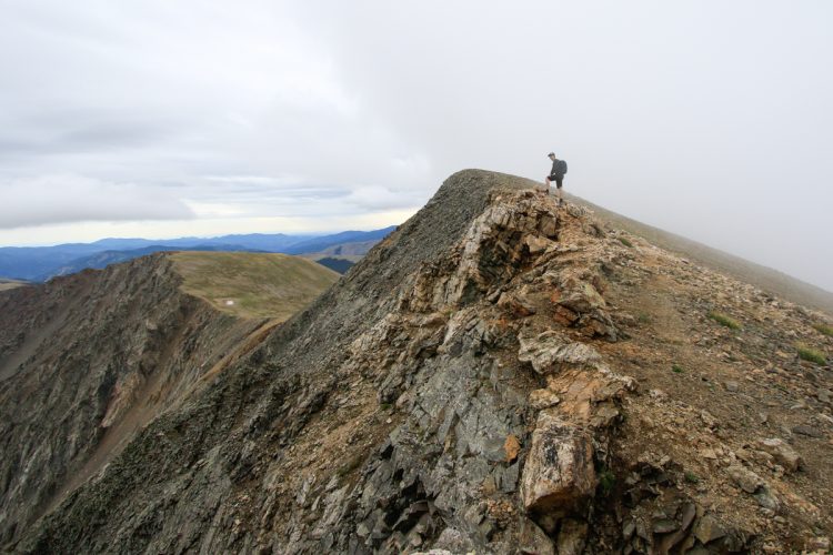 A lone hiker stands on a rocky mountain ridge, overlooking a vast landscape with rolling hills and distant mountains under a cloudy sky. The rugged terrain features dramatic cliffs and a steep drop-off on either side of the hiker, showcasing the beauty of nature in a remote location.