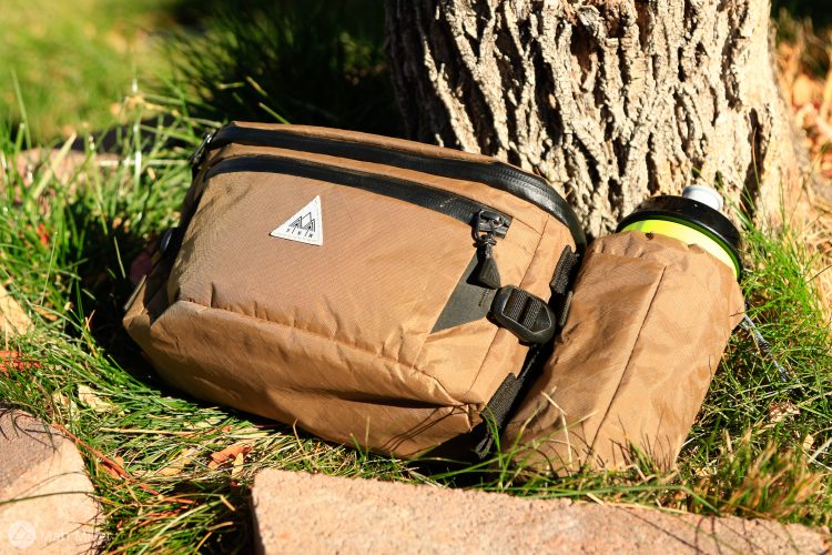 A brown, weather-resistant bag resting on the grass near a tree, with a water bottle in a sleeve attached to its side. The bag has a triangular logo and features zippers and adjustable straps. Surrounding the bag are small stones and patches of green grass.