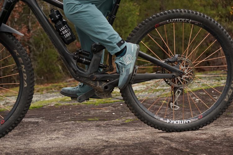 A close-up of a mountain bike in motion, highlighting the bike's frame, chain, and rear wheel with copper spokes. A rider's foot is positioned on the pedal, wearing teal cycling shoes. A water bottle labeled "Earl's Bike Shop" is attached to the bike frame, set against a natural backdrop.