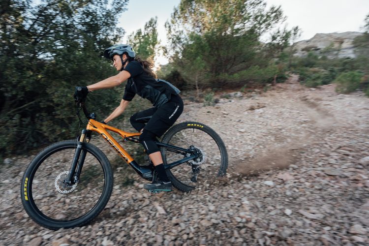 A mountain biker riding an orange bicycle on a rocky trail, surrounded by trees. The rider is wearing a helmet and sports attire, leaning forward as they maneuver the path, with dust and gravel kicking up from the rear tire.