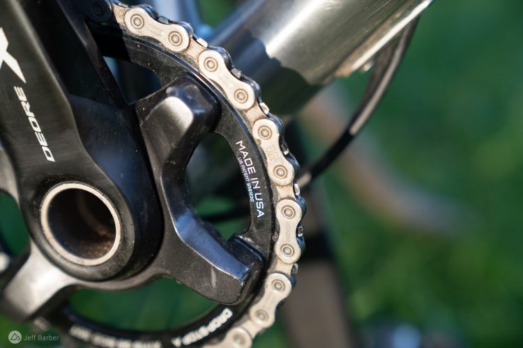 Close-up view of a bicycle's crankset and chain, featuring a Shimano Deore XT component with a sleek black finish. The chain is shown engaging with the crank teeth, highlighting the engraved "MADE IN USA" labeling. The background is softly blurred greenery, emphasizing the mechanical details of the bike part.