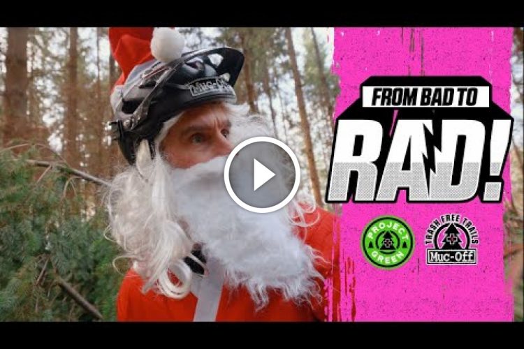 A person wearing a Santa hat and a white beard, equipped with a black helmet, surrounded by trees in a forest setting. The image includes text that reads "FROM BAD TO RAD!" along with logos for Project Green and Trash Free Trails. A play button icon indicates that the image is part of a video.