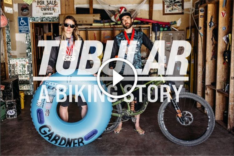 A man and a woman stand in a rustic indoor space decorated with various signs and memorabilia. The man is holding a mountain bike and wearing a black shirt, while the woman holds a blue inflatable inner tube and wears sunglasses, along with medals around her neck. The text overlay reads "TUBULAR A BIKING STORY."