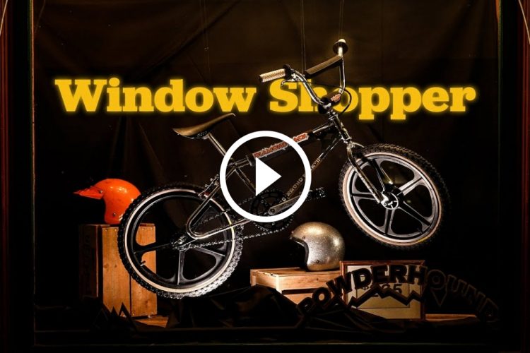 A stylized window display featuring a black BMX bike prominently suspended in front of a dark background. The words "Window Shopper" are displayed in bright yellow above the bike. In the foreground, there is an orange helmet and a metallic silver helmet, along with wooden crates and a decorative sign reading "POWDERHOUND." The display creates an eye-catching, artistic presentation of biking culture.