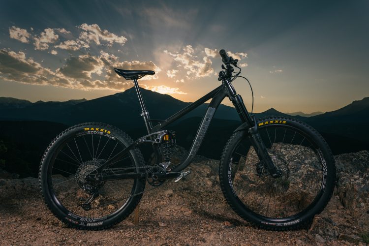 A mountain bike is positioned on a rocky surface against a backdrop of mountains and a dramatic sunset. The bike features a sleek black frame, prominent tires labeled "Maxxis," and a raised seat. The sky is filled with clouds and vibrant colors from the setting sun, highlighting the rugged terrain.
