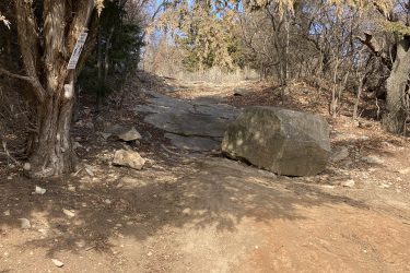 A rocky trail leading upward through a wooded area, with a large boulder on the right and a road sign attached to a tree on the left. The ground is covered in dirt and small rocks, with sparse vegetation in the background. Lake Ponca Trails mountain bike trail.