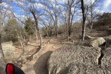A winding dirt path through a wooded area with bare trees and rocky terrain under a partly cloudy sky. The scene shows a natural setting with scattered rocks and fallen leaves, suggesting a tranquil outdoor environment. A portion of a red object is visible in the lower left corner, likely part of a hiking or biking gear. Lake Ponca Trails mountain bike trail.
