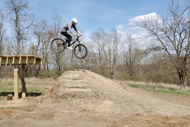 A person wearing a helmet is performing a jump on a bicycle off a wooden ramp, soaring above a dirt mound. The background features trees and a clear blue sky, indicating a sunny day. Creekside Trails mountain bike trail.