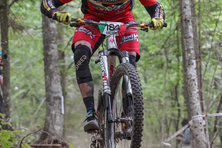 A mountain biker in a red jersey and black shorts is skillfully navigating a dirt trail surrounded by trees. The cyclist is performing a jump on their bike, with both wheels off the ground, showcasing athleticism and focus. Dust is kicking up from the trail, adding to the dynamic atmosphere of the outdoor racing event.