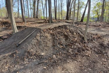 A dirt ramp or jump built in a forested area, surrounded by trees and fallen leaves, with a wooden tool resting on the ground beside it. The scene is illuminated by sunlight filtering through the branches, indicating a clear day. Creekside Trails mountain bike trail.