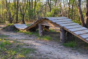 A wooden pedestrian bridge with a gently arching design, surrounded by lush greenery and trees in a forested area. A dirt path leads off to the left of the bridge, with natural vegetation peeking through the ground. The sunlight filters through the trees, creating a peaceful outdoor ambiance. Creekside Trails mountain bike trail.