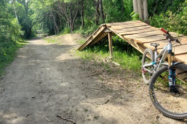 A mountain bike leaning against a wooden ramp on a dirt trail surrounded by lush green trees. The path extends into the distance, showcasing a peaceful outdoor setting ideal for cycling. Creekside Trails mountain bike trail.