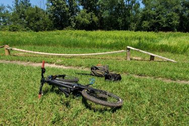 A black mountain bike lies on its side on green grass, with a wooden ramp in the background. Two black bags are placed nearby, surrounded by tall grass and trees in the distance under a clear blue sky. Creekside Trails mountain bike trail.