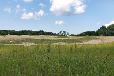 A landscape view featuring a bike or dirt jumping area with several earth mounds. The scene includes tall grasses in the foreground and a backdrop of trees under a partly cloudy sky. A wooden ramp structure is visible on the right side of the image. Creekside Trails mountain bike trail.