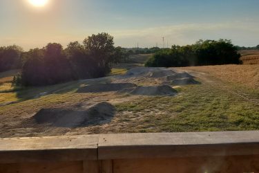 A scenic view of a dirt bike track featuring several jumps, set against a backdrop of rolling fields and trees under a clear sky with a bright sun. The ground is mostly bare with patches of grass, and the landscape features tall grass and distant hills. An elevated wooden railing is visible in the foreground. Creekside Trails mountain bike trail.