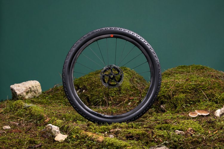 A close-up of a bicycle wheel positioned upright on a bed of moss and rocks, with a solid green background. The wheel features a black tire with a textured tread and a prominent brake disc at the center.