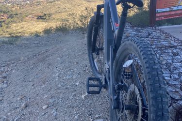 A mountain bike is parked on a rocky trail overlooking a scenic view of a valley and distant mountains. A trail sign indicating hiking information is visible in the background. The sky is partially cloudy, and the landscape features a mix of urban and natural elements. 7th St. and Mineral mountain bike trail.
