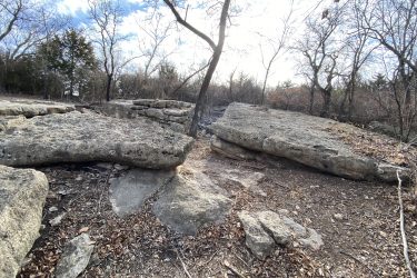 A rocky landscape featuring large boulders and scattered stones, surrounded by bare trees. The scene is illuminated by soft sunlight filtering through the clouds, creating a serene outdoor atmosphere. Leaves are scattered on the ground, indicating a late autumn or early winter setting. Lake Ponca Trails mountain bike trail.