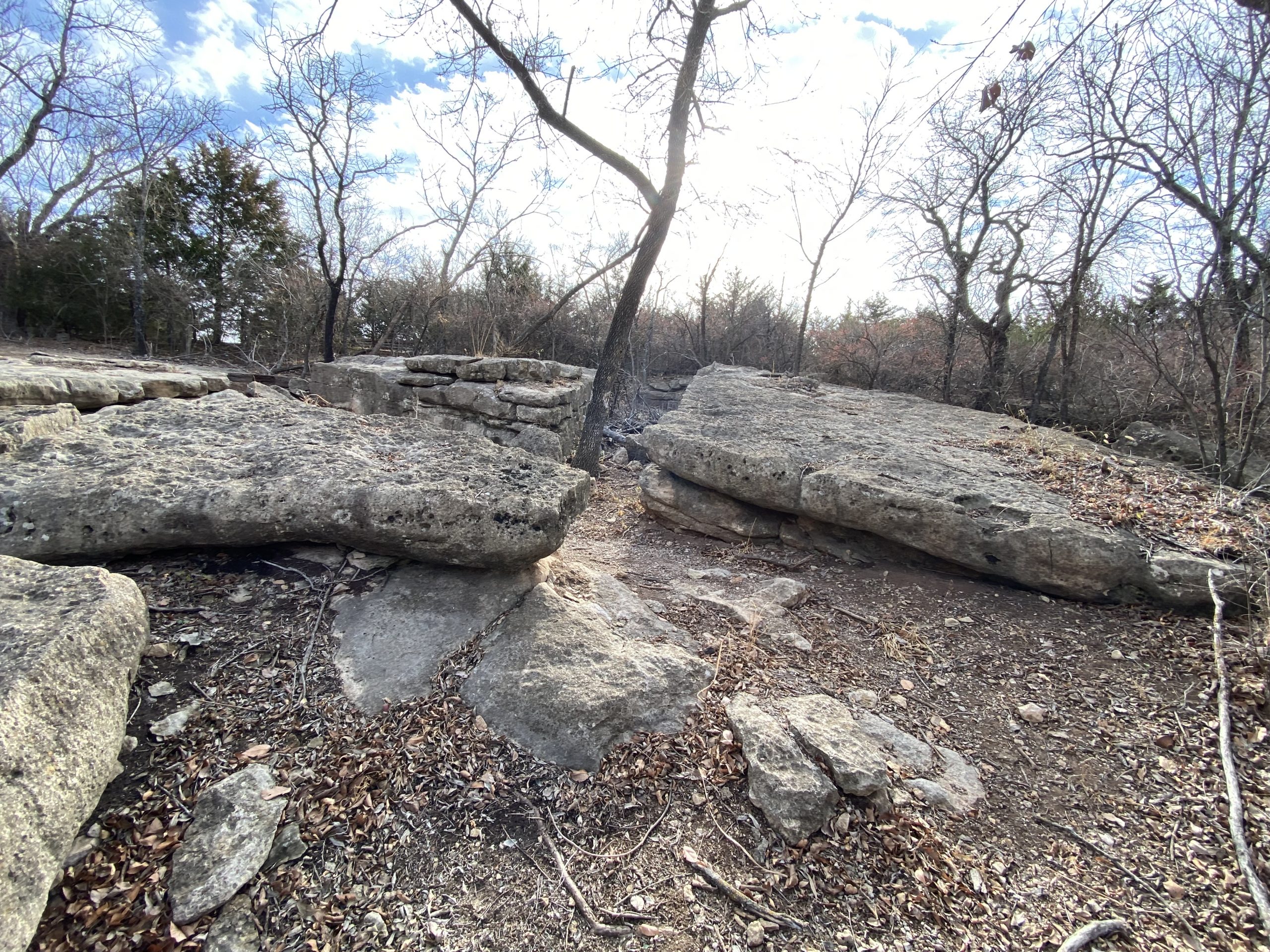A rocky landscape featuring large boulders and scattered stones, surrounded by bare trees. The scene is illuminated by soft sunlight filtering through the clouds, creating a serene outdoor atmosphere. Leaves are scattered on the ground, indicating a late autumn or early winter setting. Lake Ponca Trails mountain bike trail.