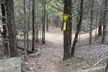 A forest trail with a sign that reads "DROP OFF" accompanied by a downward arrow, directing hikers along a winding path. The scene is surrounded by trees and natural terrain scattered with leaves and rocks. Lake Ponca Trails mountain bike trail.