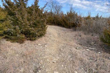 A dirt hiking trail winding through sparse vegetation with shrubs and dry grass under a partly cloudy sky. Lake Ponca Trails mountain bike trail.