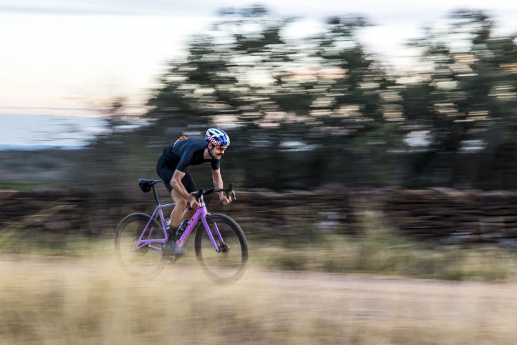 A cyclist in a black outfit and helmet rides a purple bicycle at high speed on a dirt path surrounded by grass, with trees blurred in the background to emphasize motion.