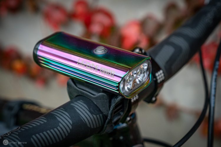 Close-up image of a vibrant, rainbow-colored bike light mounted on handlebars. The light features three bright LEDs and is labeled "LEZYNE HLM 1000XL." The handlebars have textured grips, and colorful foliage is blurred in the background, suggesting an outdoor setting.