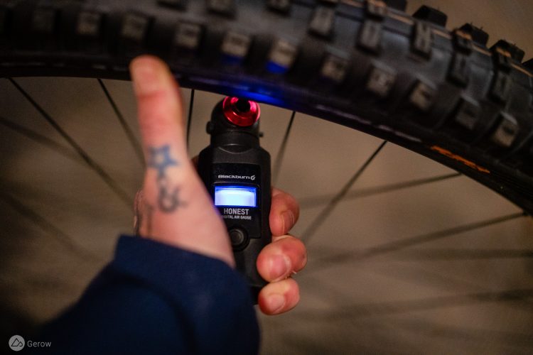 A hand holding a digital air gauge, positioned against the valve of a bicycle tire. The gauge displays a blue backlight with the word "HONEST" on it. The tire has a textured surface with visible tread patterns, and the background is softly blurred, suggesting a dimly lit environment. A tattoo is visible on the hand.