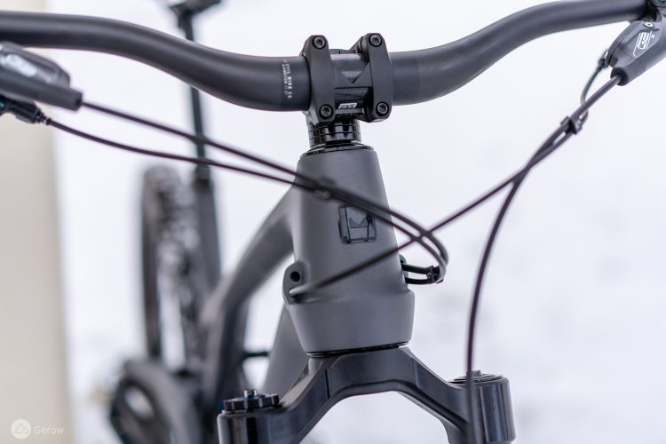 Close-up view of a bicycle's handlebar, headset, and stem, showcasing a matte black finish and intricate cable routing. The background is soft and blurred, providing focus on the bike components.