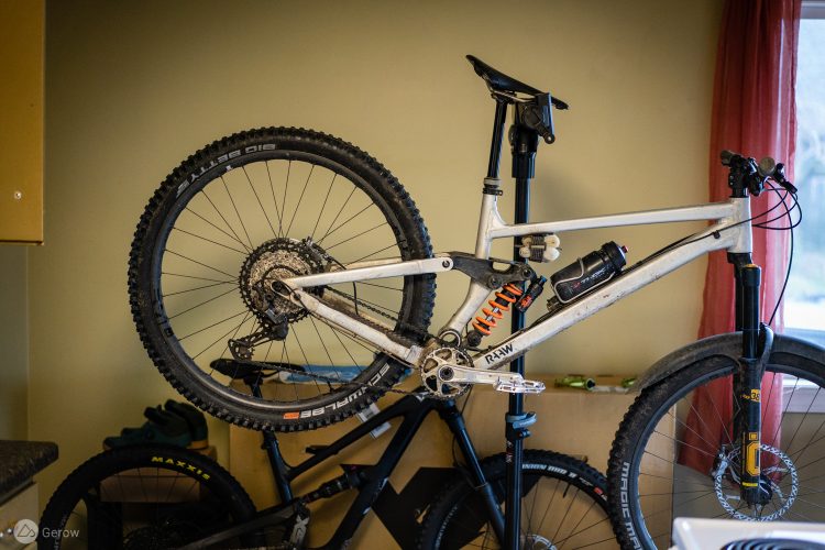A mountain bike propped against a wall, showcasing its white frame, large tires, and various components like a shock absorber and water bottle holder. The background features a neutral-colored wall and a window with a sheer curtain, along with another bike visible in the lower part of the image.