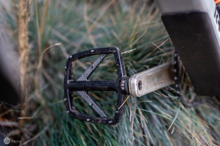 Close-up of a bicycle pedal resting on grass, showcasing the textured surface and silver crank arm. The pedal features a black frame with a geometric design and is partially obscured by blades of grass.