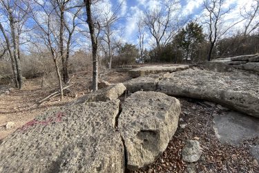 A rocky landscape featuring large boulders scattered on the ground, surrounded by bare trees and sparse vegetation. The sky is partly cloudy, creating a bright atmosphere. Fallen leaves cover the ground, adding to the natural setting. Lake Ponca Trails mountain bike trail.