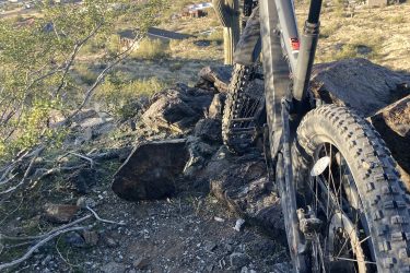 A mountain bike is positioned on rocky terrain with a view of a desert landscape below, featuring scattered homes and vegetation. In the background, cacti and distant hills are visible under a clear sky. 7th St. and Mineral mountain bike trail.