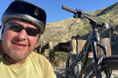 A person wearing a black helmet and sunglasses smiles for a selfie next to a mountain bike. The background features a mountainous landscape with clear blue skies. 7th St. and Mineral mountain bike trail.