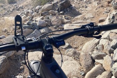 Close-up view of a mountain bike's handlebars on a rocky trail, with a blurred background of stones and sparse vegetation. The perspective suggests the rider is preparing to navigate a challenging terrain. 7th St. and Mineral mountain bike trail.
