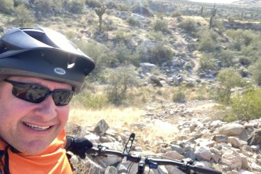 A person wearing a black bicycle helmet and sunglasses smiles while holding the handlebars of a mountain bike. The background features a rocky landscape with desert vegetation, including cacti and rolling hills under bright sunlight. 7th St. and Mineral mountain bike trail.