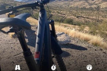 A mountain bike parked on a gravel trail with a scenic view of mountains and desert landscape in the background. The display shows statistics: 11.92 miles distance, 1 hour and 17 minutes duration, and an average speed of 9.2 mph. 7th St. and Mineral mountain bike trail.