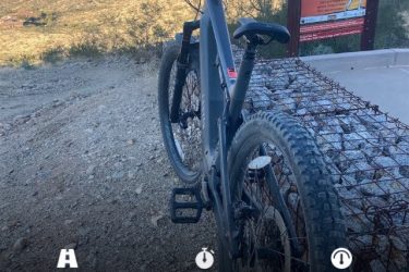 A mountain bike parked on rocky terrain, with a view of a distant cityscape under clear skies. Overlay text indicates a distance of 14.77 miles, a duration of 1 hour and 17 minutes, and an average speed of 11.5 mph. A heat warning sign is partially visible in the background. 7th St. and Mineral mountain bike trail.