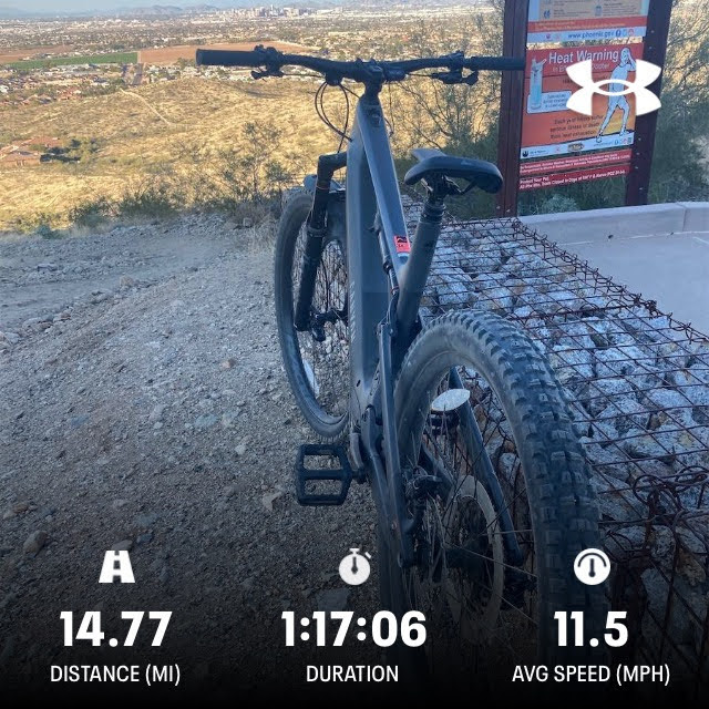 A mountain bike parked on rocky terrain, with a view of a distant cityscape under clear skies. Overlay text indicates a distance of 14.77 miles, a duration of 1 hour and 17 minutes, and an average speed of 11.5 mph. A heat warning sign is partially visible in the background. 7th St. and Mineral mountain bike trail.