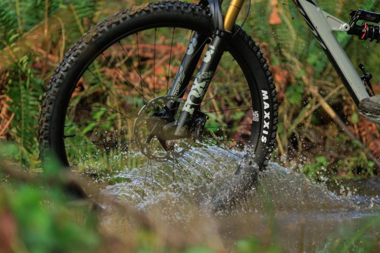 A close-up image of a mountain bike wheel splashing through water while navigating a muddy trail, surrounded by lush greenery.