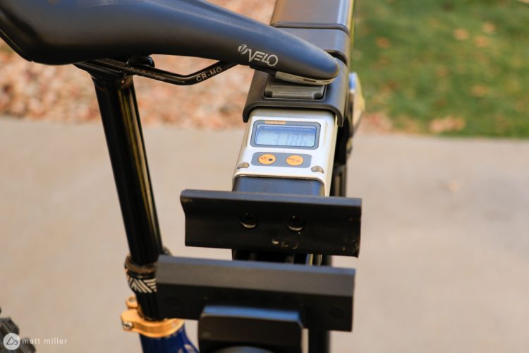 A close-up view of a bicycle saddle and a digital scale mounted on the bike frame, displaying a weight reading. The background features a blurred grassy area.
