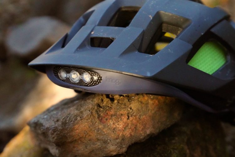 Close-up of a bicycle helmet resting on a rock, featuring a built-in LED light on the front. The helmet has a dark gray color with ventilation holes and a hint of bright green visible inside. The light has three LEDs set within a dotted black casing. Natural outdoor setting in the background.