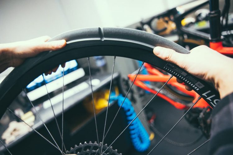 Close-up of hands holding a bicycle wheel rim in a workshop setting, with tools and a bike partially visible in the background.