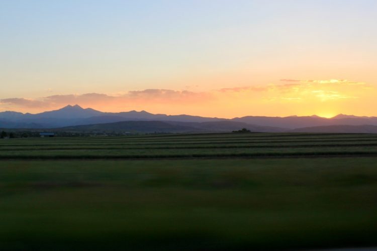 A scenic view of mountains silhouetted against a vibrant sunset, with lush green fields in the foreground. The sky displays hues of orange, pink, and blue, creating a tranquil atmosphere.